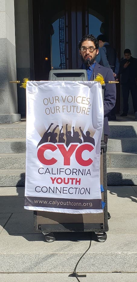 Ricardo standing in front of a podium giving a speech at shadow day in Sacramento, California. He is wearing a suit and tie and is speaking to an audience about the importance of supporting disabled foster youth.